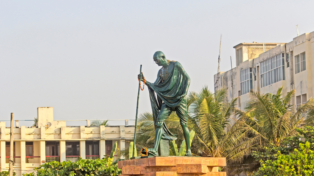 Statue of Mahatma Gandhi wearing round spectacles and a draped shawl, holding a walking stick with one hand raised.